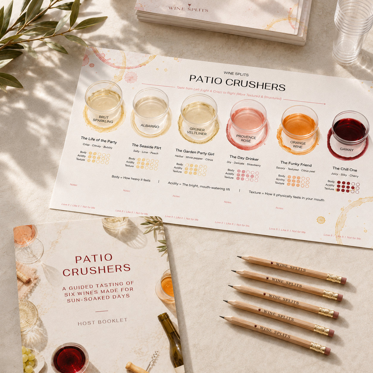 Wine Splits Patio Crushers kit layout: tasting placemat with six wine pours, host booklet, and branded pencils on a linen surface